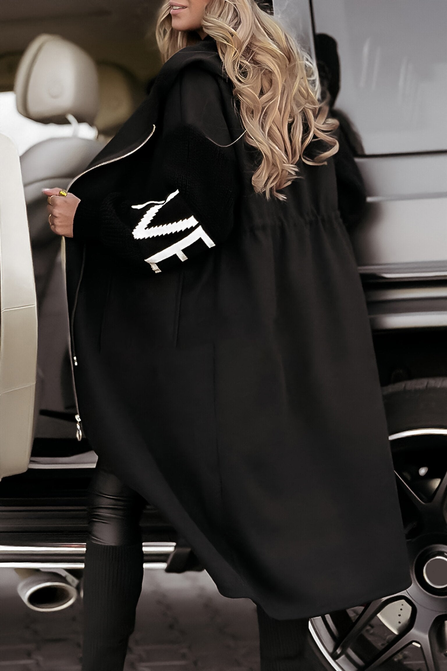 Person wearing a black coat with a logo, standing next to a car.