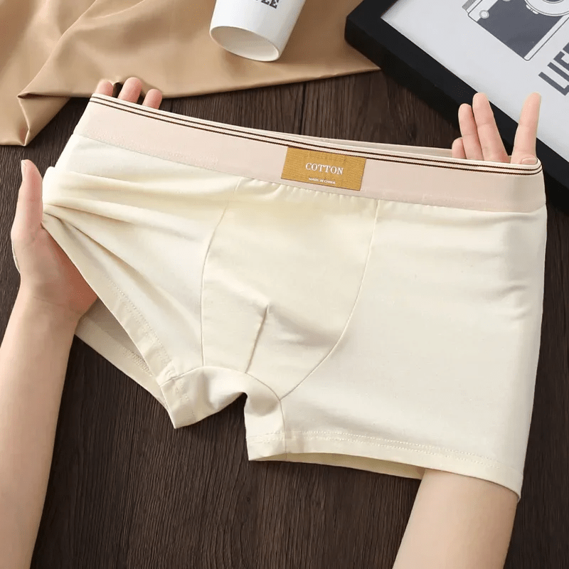 Beige underwear held up on a wooden surface with a cup and fabric in the background.