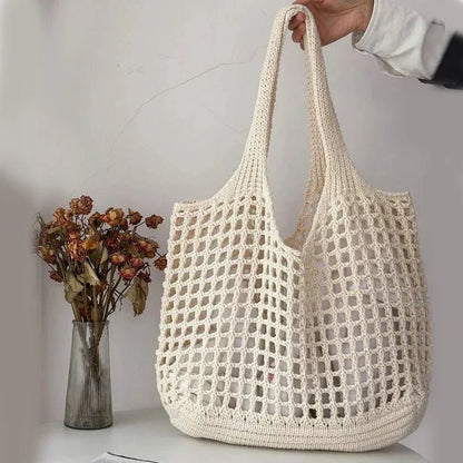 Beige woven bag held by a hand with a vase of dried flowers in the background.