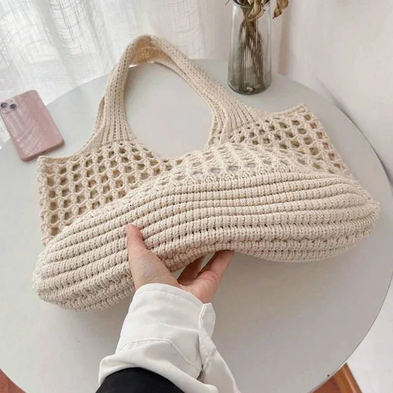 Beige woven handbag held by a person on a white surface with a phone and vase in the background.
