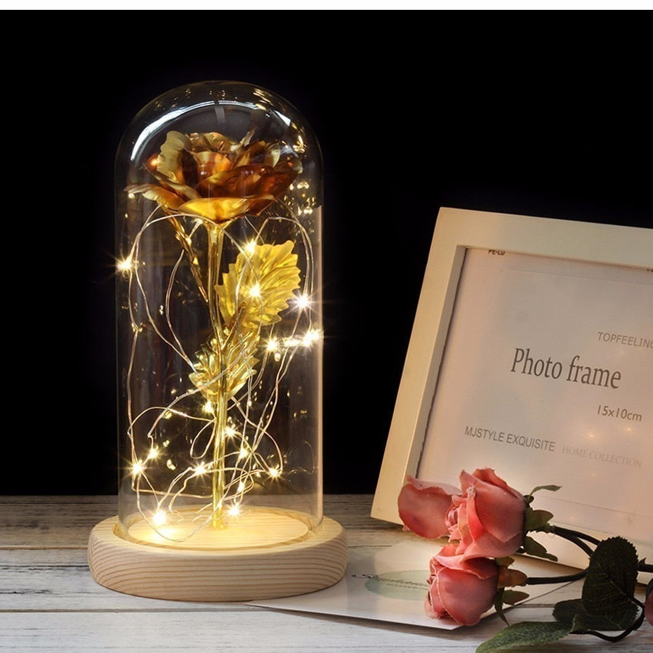 Gold rose inside a glass dome with LED lights on a wooden surface, next to a photo frame.