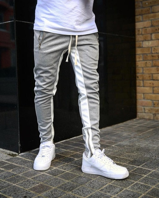 Person wearing gray jogger pants with white stripes and white sneakers in front of a black and brick wall.