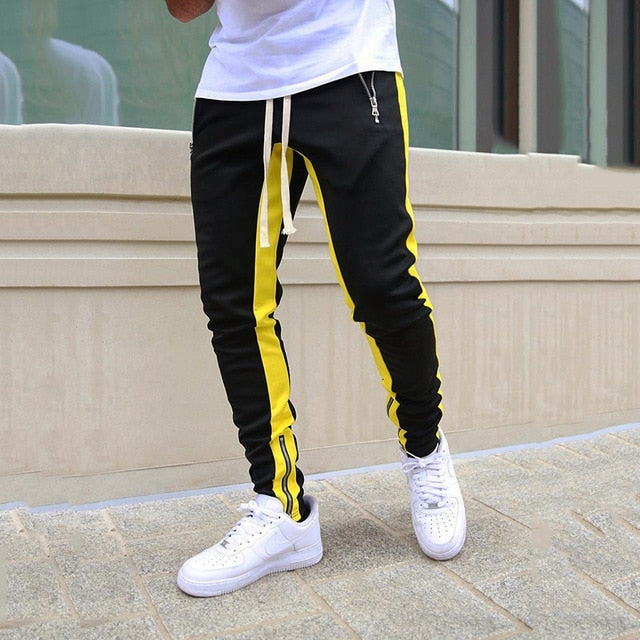 Person wearing black jogger pants with yellow stripes and white sneakers on a pavement.