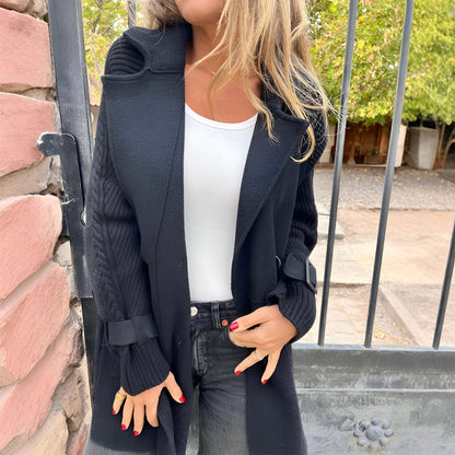 Person wearing a long dark coat over a white top and jeans, standing outdoors.