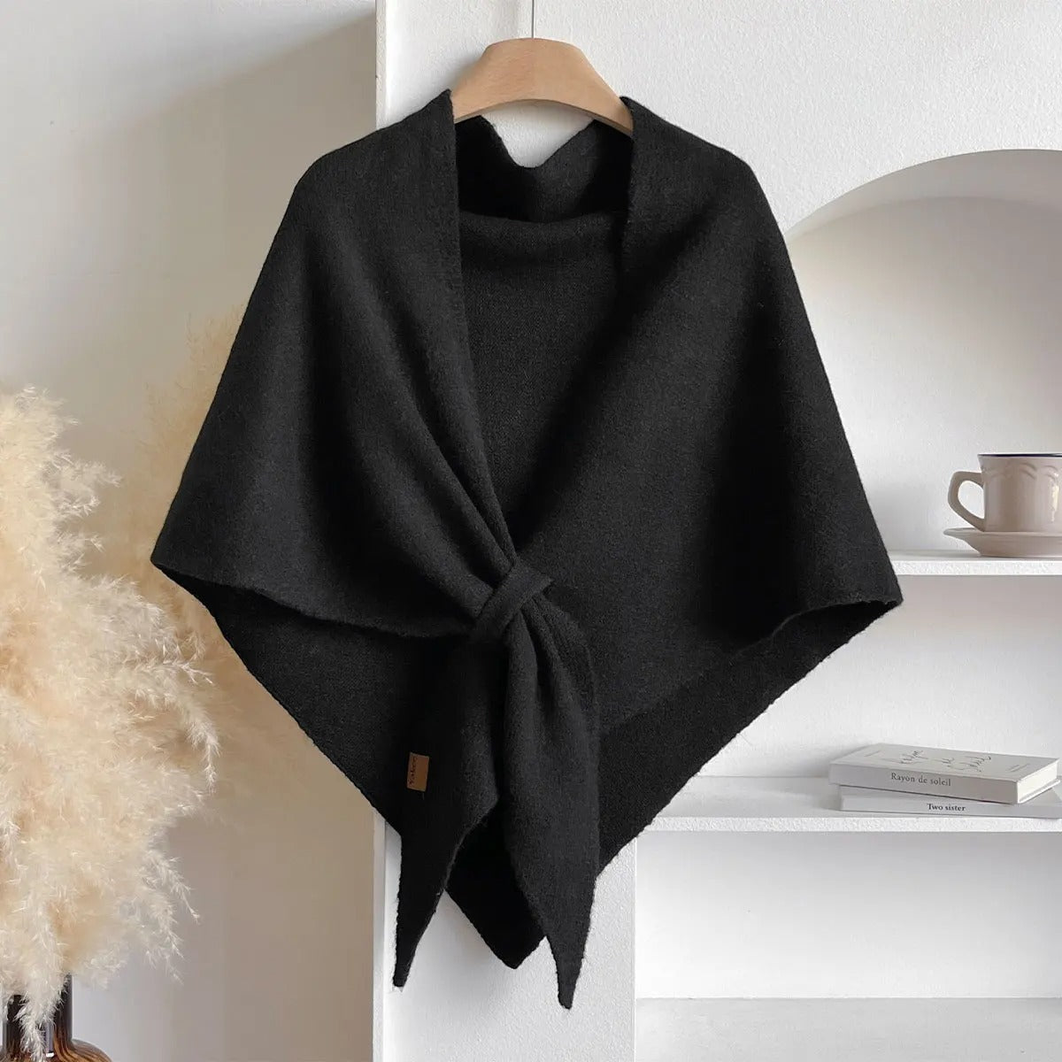 Black poncho on a hanger against a white wall with a shelf in the background.
