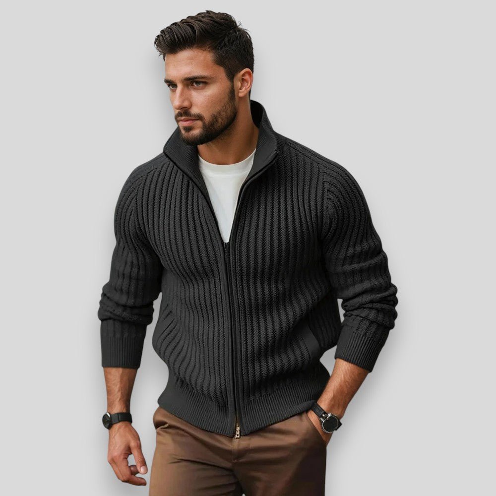 Man wearing a dark gray zip-up sweater over a white shirt with brown pants on a light gray background