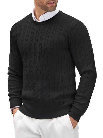 Man wearing a black cable knit sweater over a white shirt with white pants on a white background